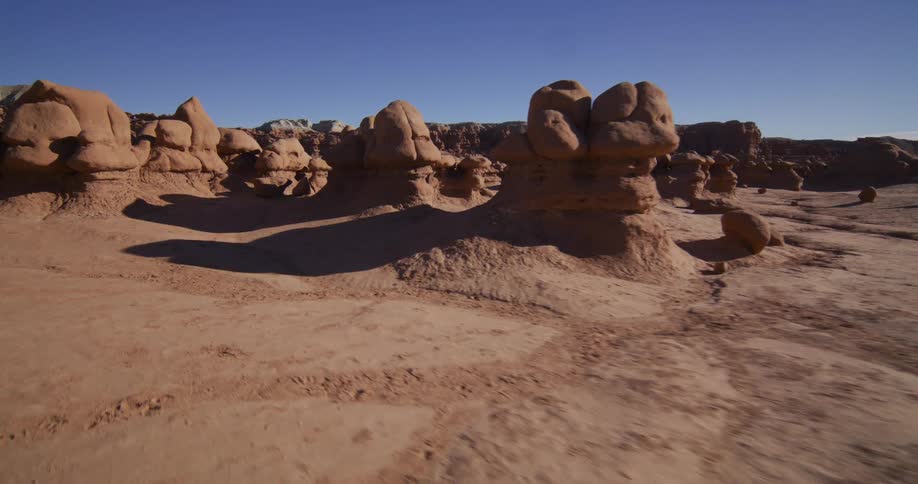A slow and cinematic aerial tracking shot flies through the otherworldly landscape of Goblin Valley, Utah, revealing its unique mushroom-shaped hoodoo rock formations under a clear blue sky