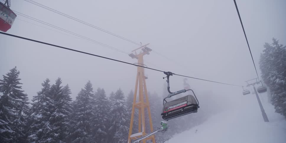 A ski lift and gondola pass by in slow motion on a snowy and foggy day at a mountain resort