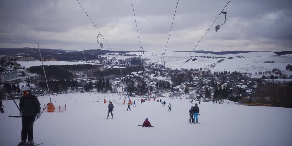 A scenic view from a t-bar lift ascending a snowy mountain, overlooking a bustling ski slope and a charming winter village