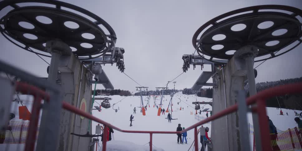 A busy ski resort with operating ski lifts and many skiers on the snowy slopes, viewed from the top of the lift machinery