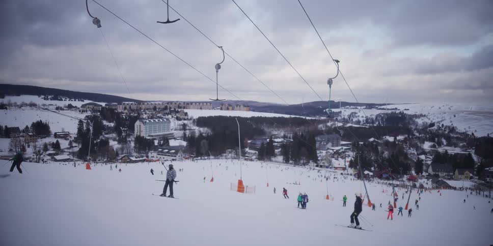Slow motion wide shot of skiers and snowboarders being pulled up a snowy slope on a T-bar lift at a bustling mountain resort