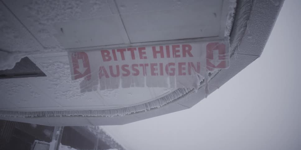A frozen German sign that says "Please get off here" is covered in thick ice and frost during a blizzard at a mountain ski resort