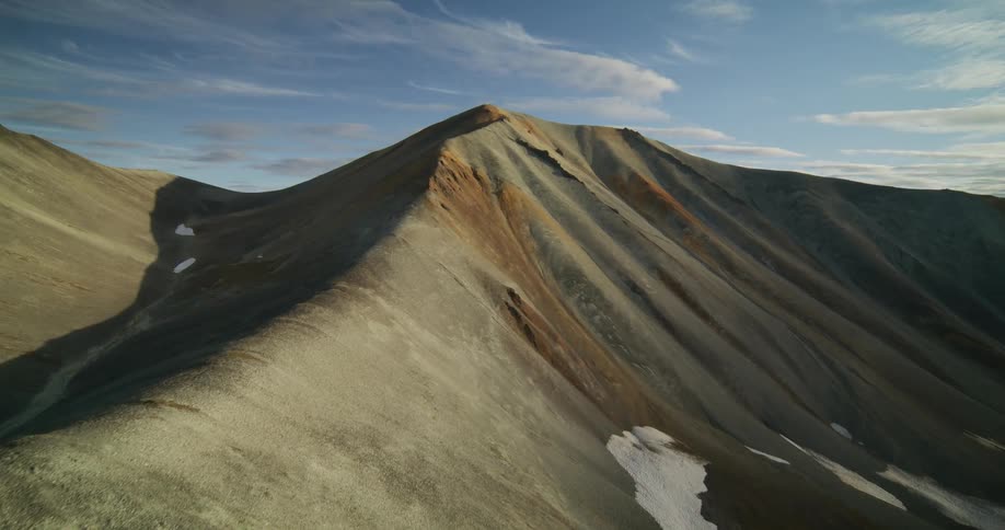 An aerial dolly shot moving towards a colorful, rugged mountain peak at golden hour