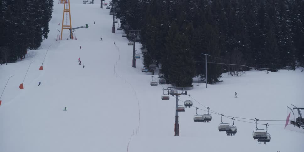 A wide view of a snowy ski resort shows skiers descending a slope next to a moving chairlift full of people