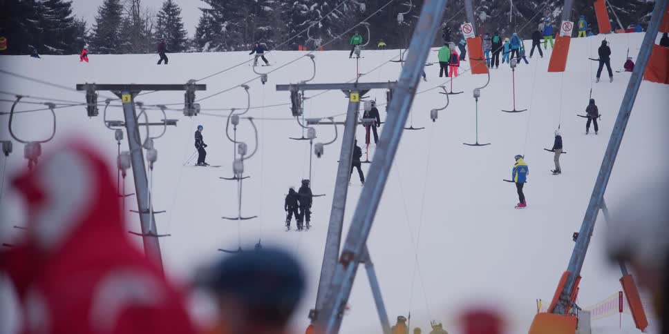 Long shot of numerous skiers being pulled up a snowy mountain slope by a T-bar lift system at a busy resort