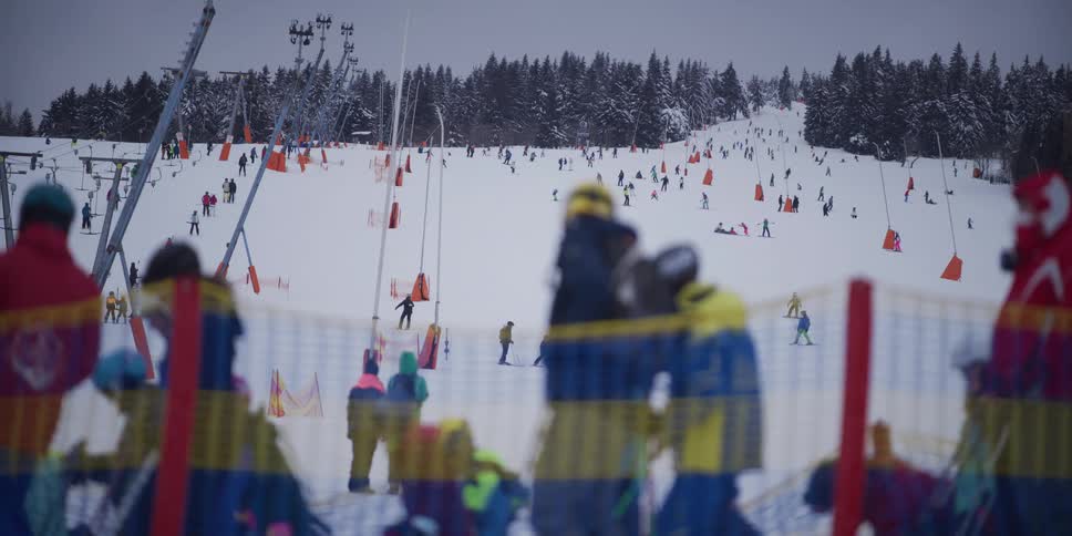 A wide, static view of a bustling ski resort slope filled with numerous skiers and snowboarders on an overcast winter day