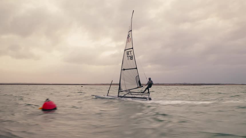 A lone sailor skillfully navigates a high-performance sailing dinghy across choppy waters under a dramatic, cloudy sky