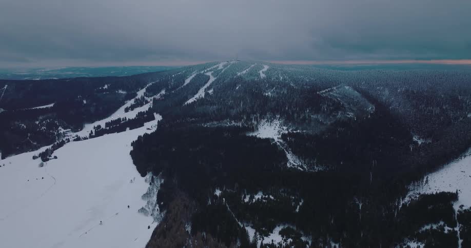 A stationary aerial shot captures the vast, dramatic landscape of a snowy mountain range with dense forests and ski slopes at twilight