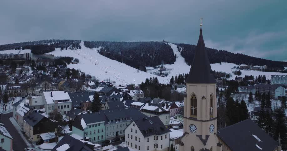 Aerial view pushing in on a historic church in a snowy mountain town with a bustling ski resort in the background at dusk