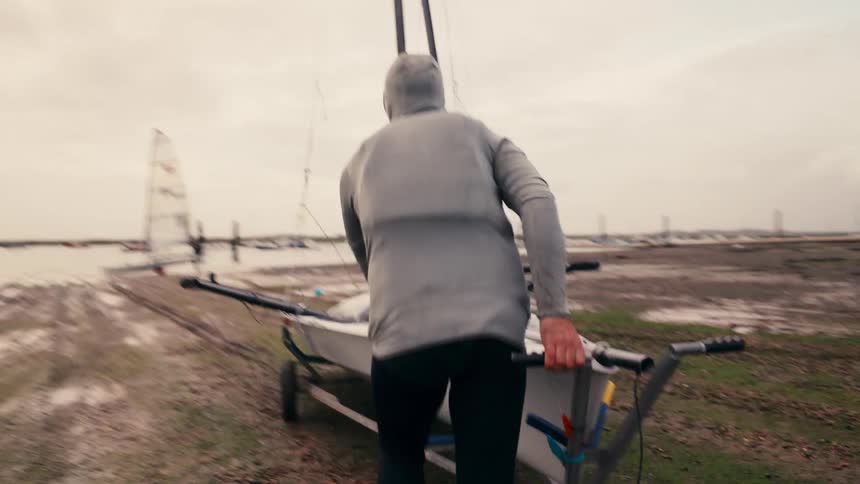 A sailor in a hooded wetsuit pushes his small racing dinghy on a dolly across the muddy shore towards the water to go sailing