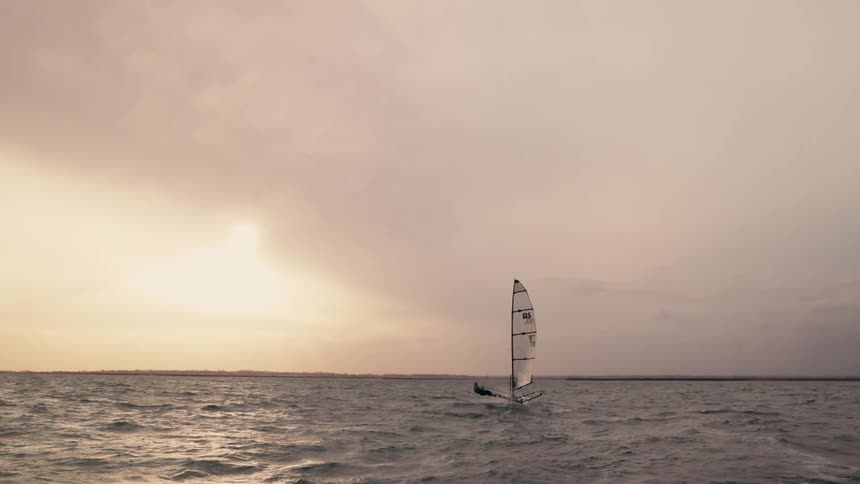 A lone sailboat navigates the choppy ocean waves under a dramatic, hazy sunset sky