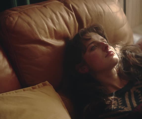 A pensive young woman rests on a couch, slowly opening her eyes as sunlight falls on her face