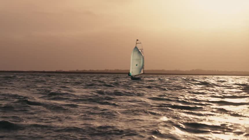 A lone sailor maneuvers a small catamaran across the choppy sea during a beautiful golden sunset