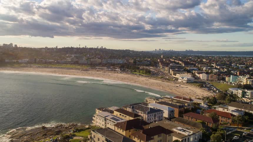 An aerial drone shot rises over rocky cliffs to reveal Sydney's famous Bondi Beach and the city skyline during golden hour
