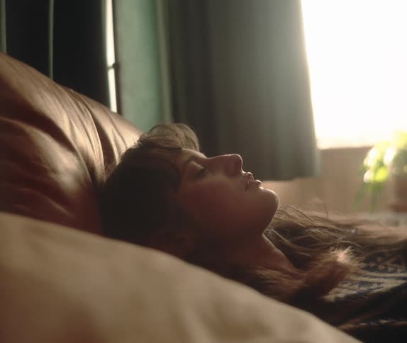 A young woman with long hair lies in bed in a sunlit room and slowly closes her eyes to rest