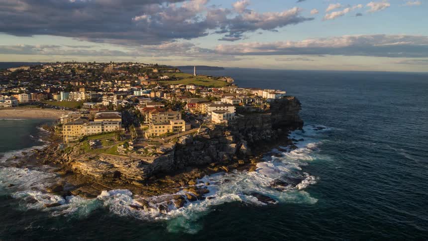 Sweeping aerial footage captures the beautiful Bondi Beach coastline in Sydney, Australia, with the city illuminated by the golden light of sunrise