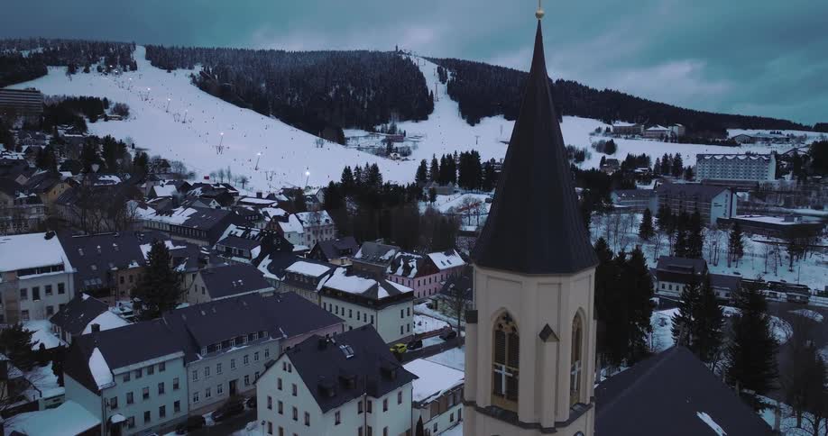 An aerial view rises above a historic church clock tower in a quaint snowy town at the base of a bustling ski resort mountain at dusk