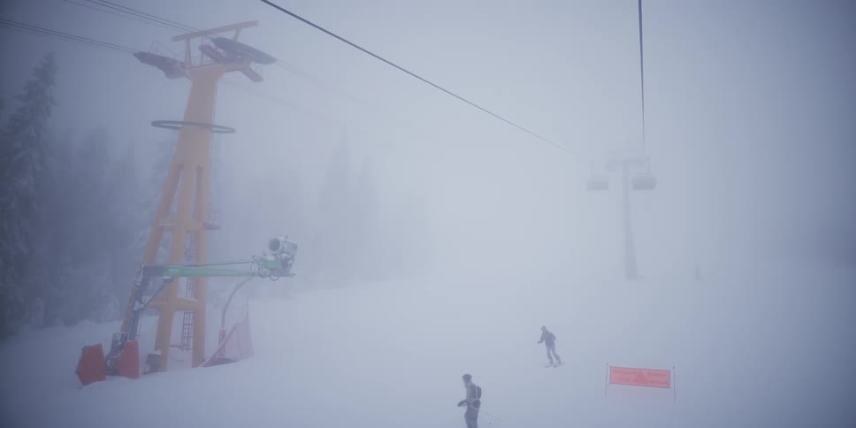 A ski lift moves slowly through the dense fog above skiers on a snowy mountain
