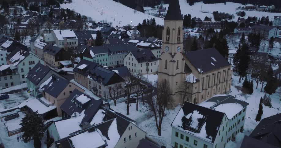 Aerial view tilts up from the snow-covered rooftops of a quaint mountain village to reveal a historic church and a bustling ski resort on the slopes beyond at dusk