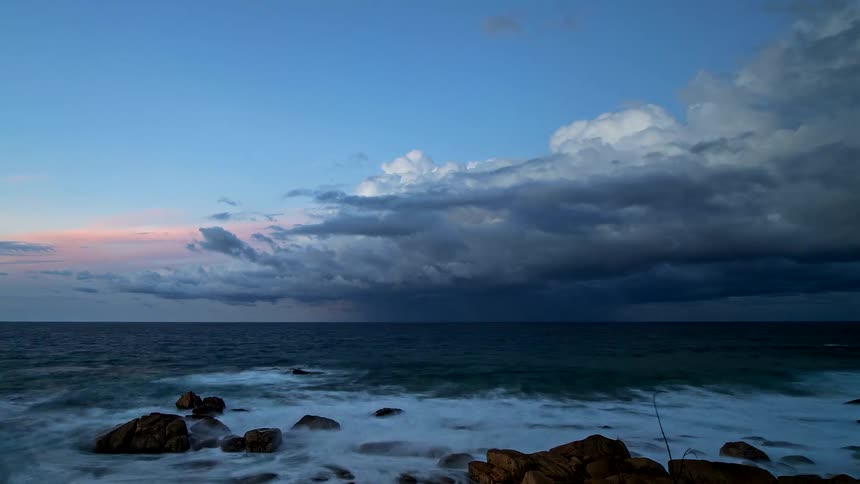 A dramatic timelapse shows a powerful storm with dark clouds rolling in over the ocean as waves crash on the rocky shore at sunset