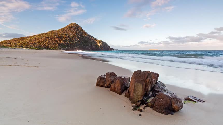A serene timelapse of ocean waves washing ashore on a sandy beach with a prominent headland, as the day transitions to a colorful sunset
