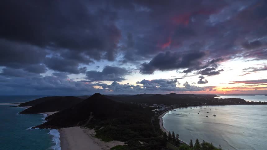A dramatic time-lapse captures a fiery sunset with colorful clouds rolling over a coastal peninsula and bay