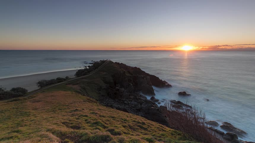 A beautiful timelapse shows the sun rising over a tranquil ocean, illuminating a grassy headland and rocky coastline