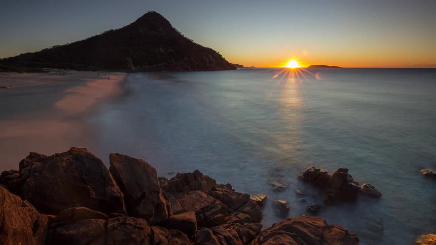 A serene time-lapse shows the sun rising over a tranquil ocean, illuminating a rocky coastline and a sandy beach beside a majestic mountain