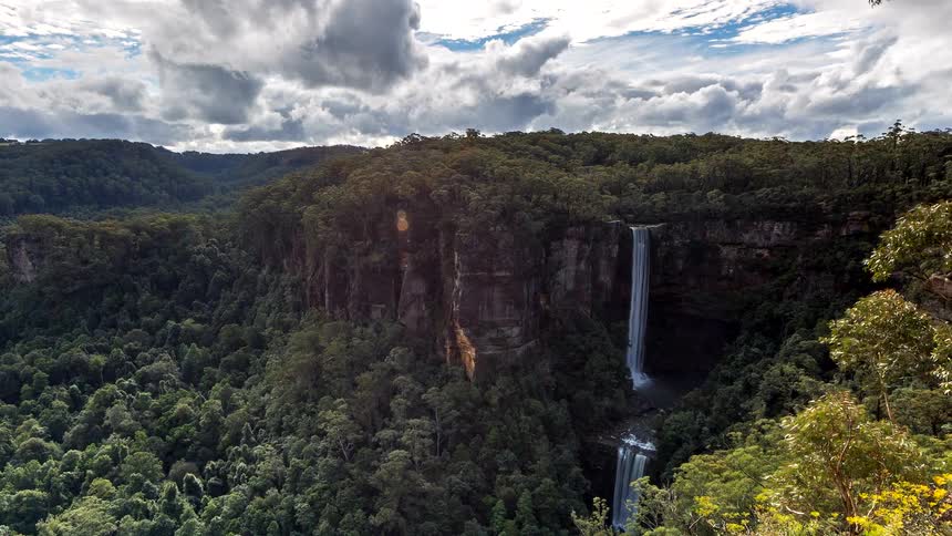 A majestic waterfall cascades down a sheer cliff into a lush, dense forest valley as dramatic clouds move quickly across the sky in a stunning time-lapse
