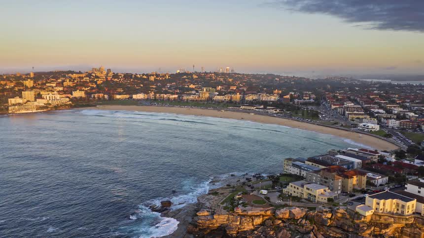 A beautiful aerial shot soars over the rocky coastline towards Sydney's iconic Bondi Beach during a golden sunrise