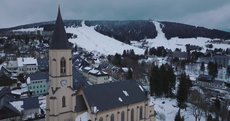 A scenic aerial view of a historic church in a snow-covered village nestled at the foot of a mountain ski resort