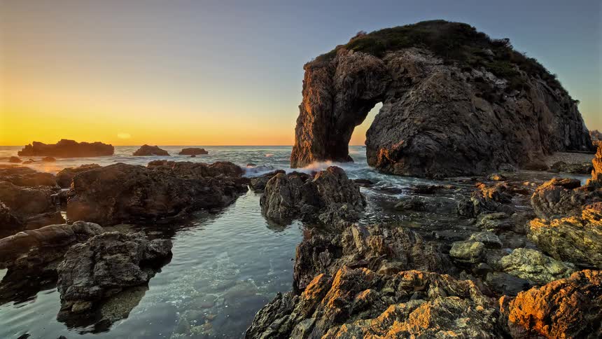 A stunning timelapse of a sunrise over a rocky coastline featuring a majestic sea arch and a calm tide pool in the foreground