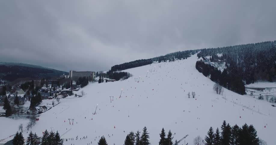 An aerial view captures the lively atmosphere of a snow-covered ski resort as skiers descend the mountain slopes under an overcast sky