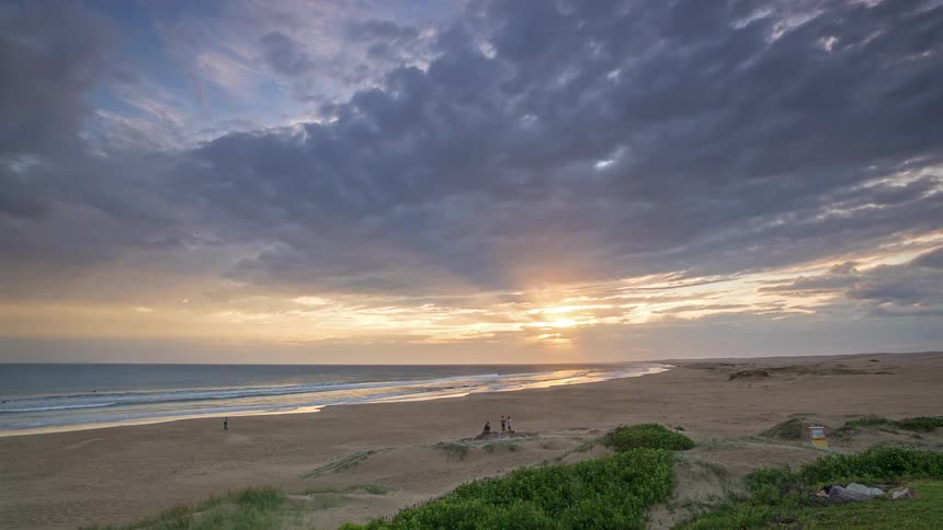 A dramatic timelapse captures the sunset over a wide sandy beach as heavy clouds roll across the sky