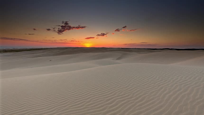 A stunning time-lapse captures a vibrant sunset over vast, rolling sand dunes as the sun disappears below the horizon and the sky turns to twilight