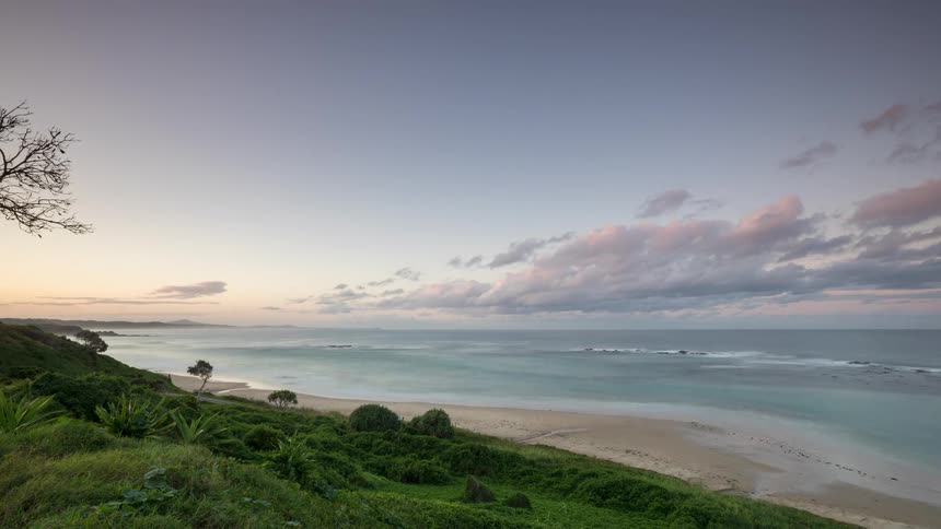 A serene time-lapse captures the transition from a beautiful sunset to a purple dusk over a calm ocean and a sandy beach from a grassy overlook