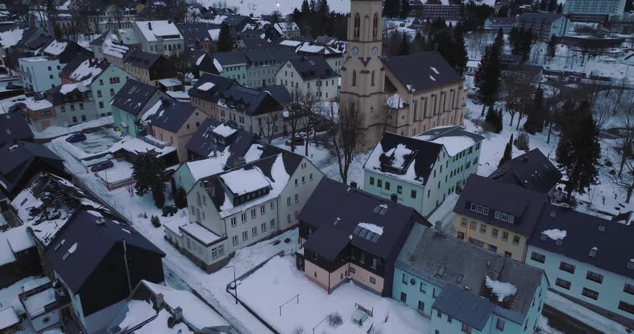 Aerial drone shot pans up to reveal a picturesque snow-covered German village with a church, nestled at the base of a mountain ski resort