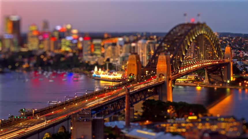 A miniature-style tilt-shift time-lapse captures streaks of light from traffic crossing the Sydney Harbour Bridge at night with the city skyline in the background