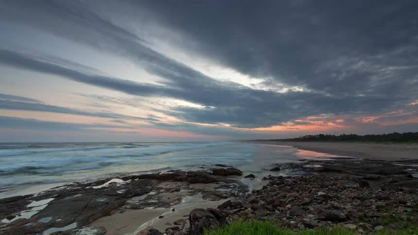 A breathtaking timelapse captures the sun setting over a rocky ocean coastline as clouds drift across the colorful evening sky