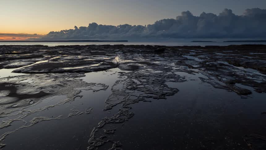 A dramatic timelapse shows clouds moving across the sky at sunrise, reflected in the still tide pools of a rocky shoreline
