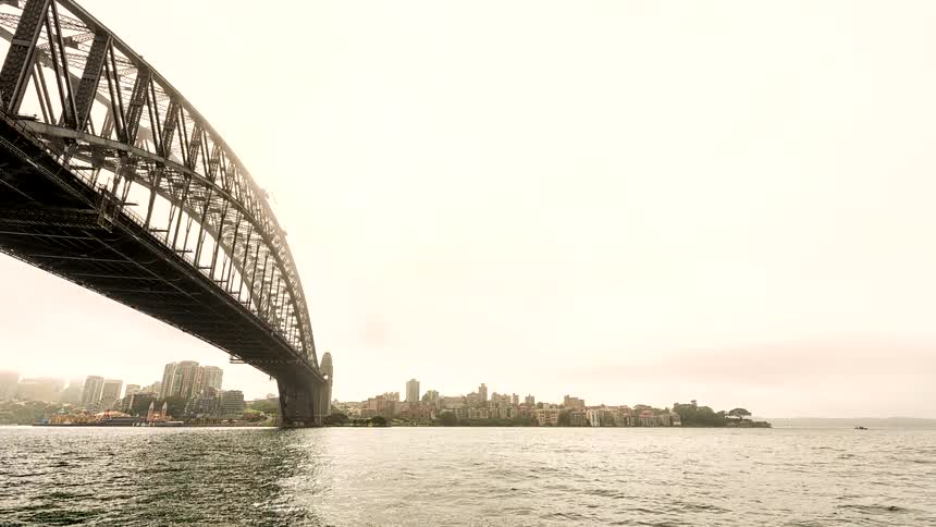 A dynamic timelapse pans from underneath the iconic Sydney Harbour Bridge, revealing the Sydney Opera House and the busy harbour on a cloudy day
