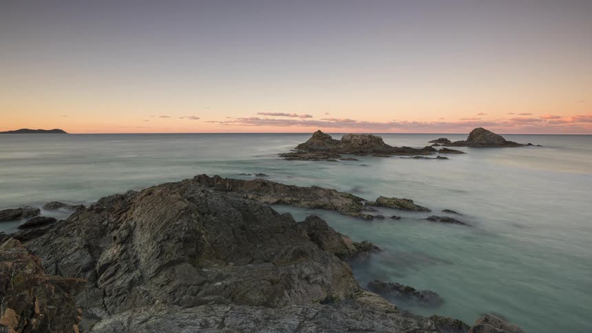 A serene long-exposure timelapse captures the vibrant colors of sunset and twilight over a rugged, rocky coastline as the ocean water flows smoothly around the shore