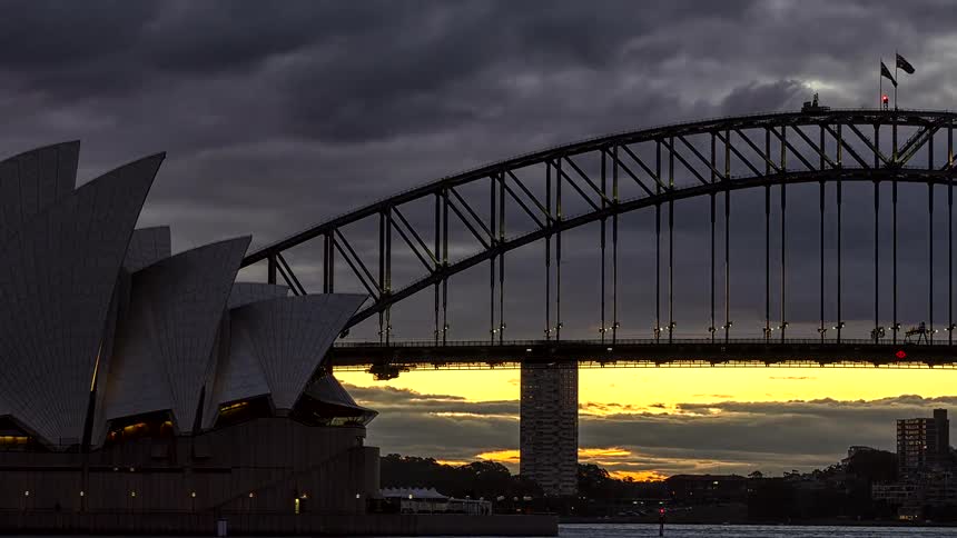 A stunning day-to-night timelapse shows a dramatic golden sunset over the iconic Sydney Opera House and Harbour Bridge as city lights turn on