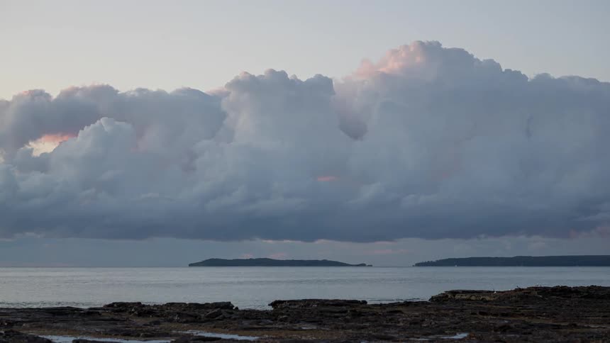 A stunning timelapse shows dramatic clouds changing colors at sunset over a calm ocean and rocky shoreline
