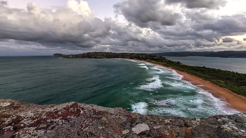 A dramatic day-to-night timelapse from a rocky clifftop shows waves crashing on a sandy peninsula as clouds sweep across the sky and lights illuminate the town