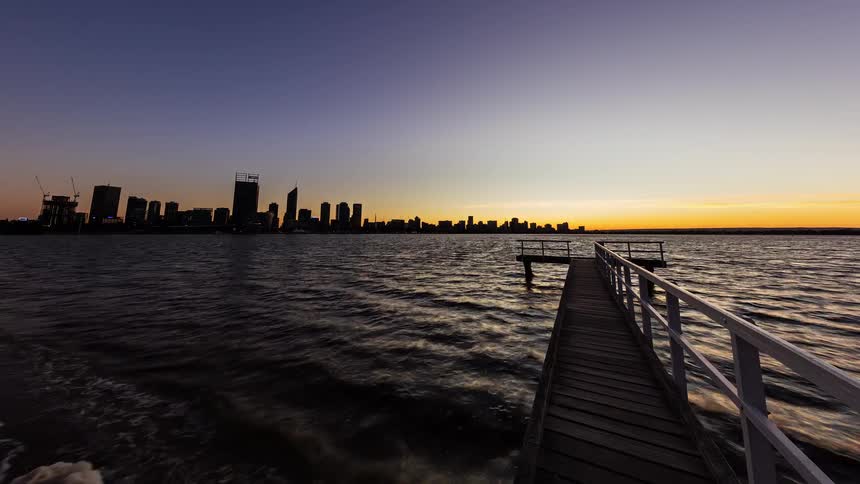 Timelapse from a wooden pier shows the spectacular transition from a glowing city skyline at night to a brilliant sunrise over the water