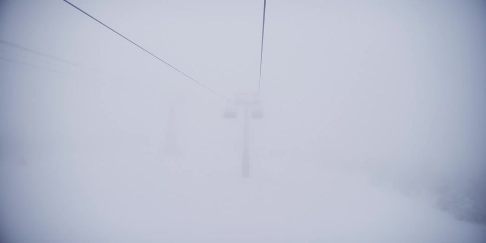 A slow-motion point-of-view shot from a chairlift ascending a snowy mountain through a heavy fog