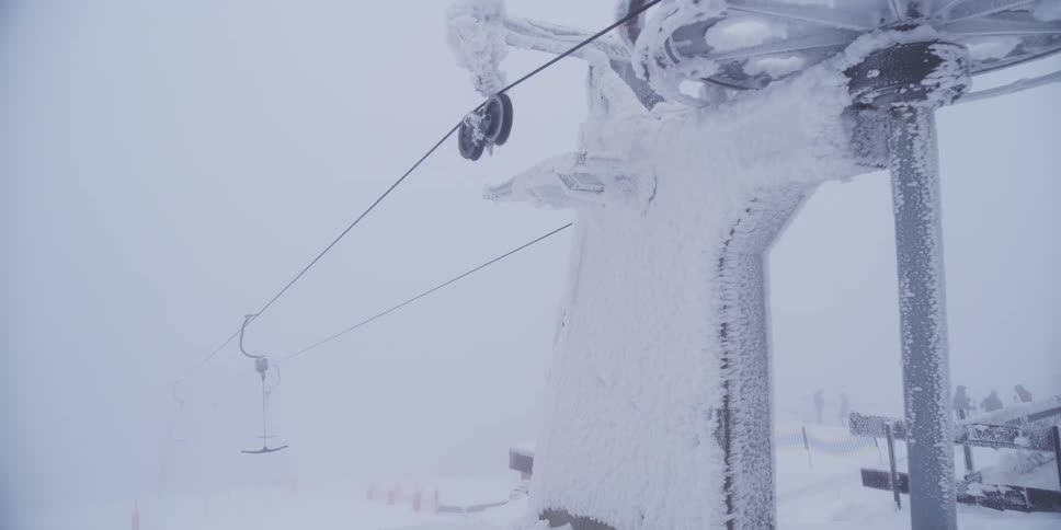 A frozen T-bar ski lift operates slowly, covered in ice and snow during a heavy blizzard on a mountain