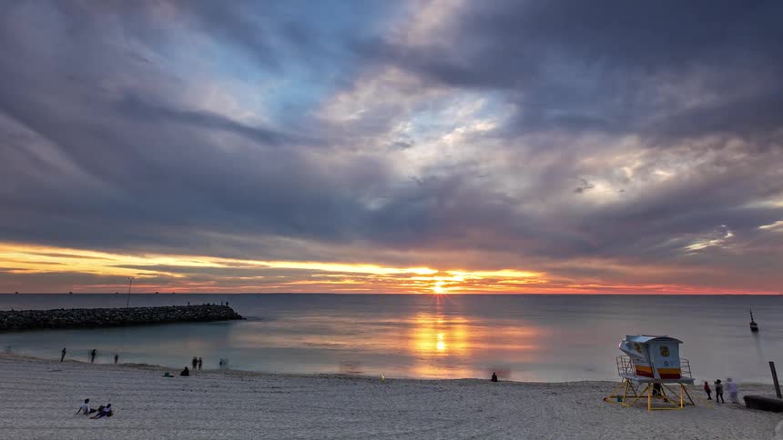 A stunning timelapse of a vibrant sunset over the ocean, with dramatic clouds moving across the sky above a sandy beach and a lifeguard tower
