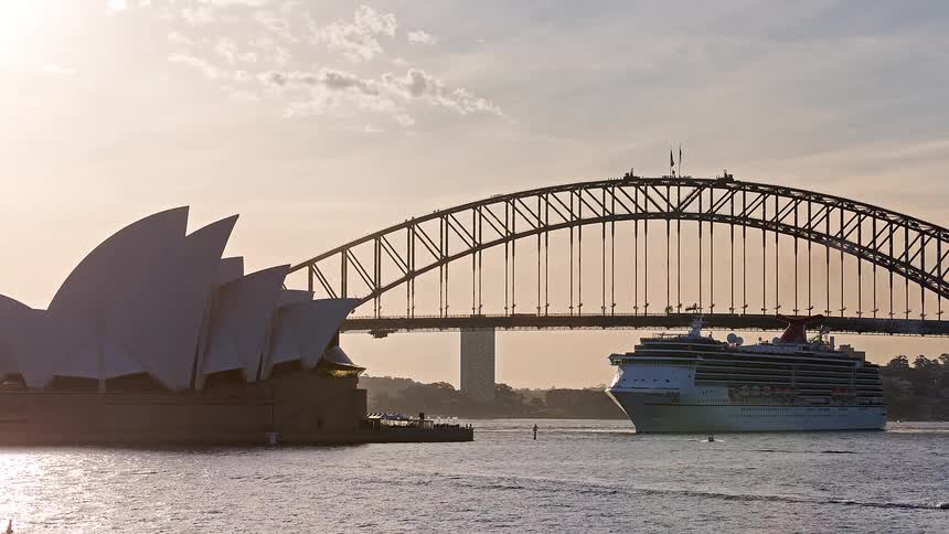 A large cruise ship sails past the Sydney Opera House and under the Harbour Bridge during a golden sunset